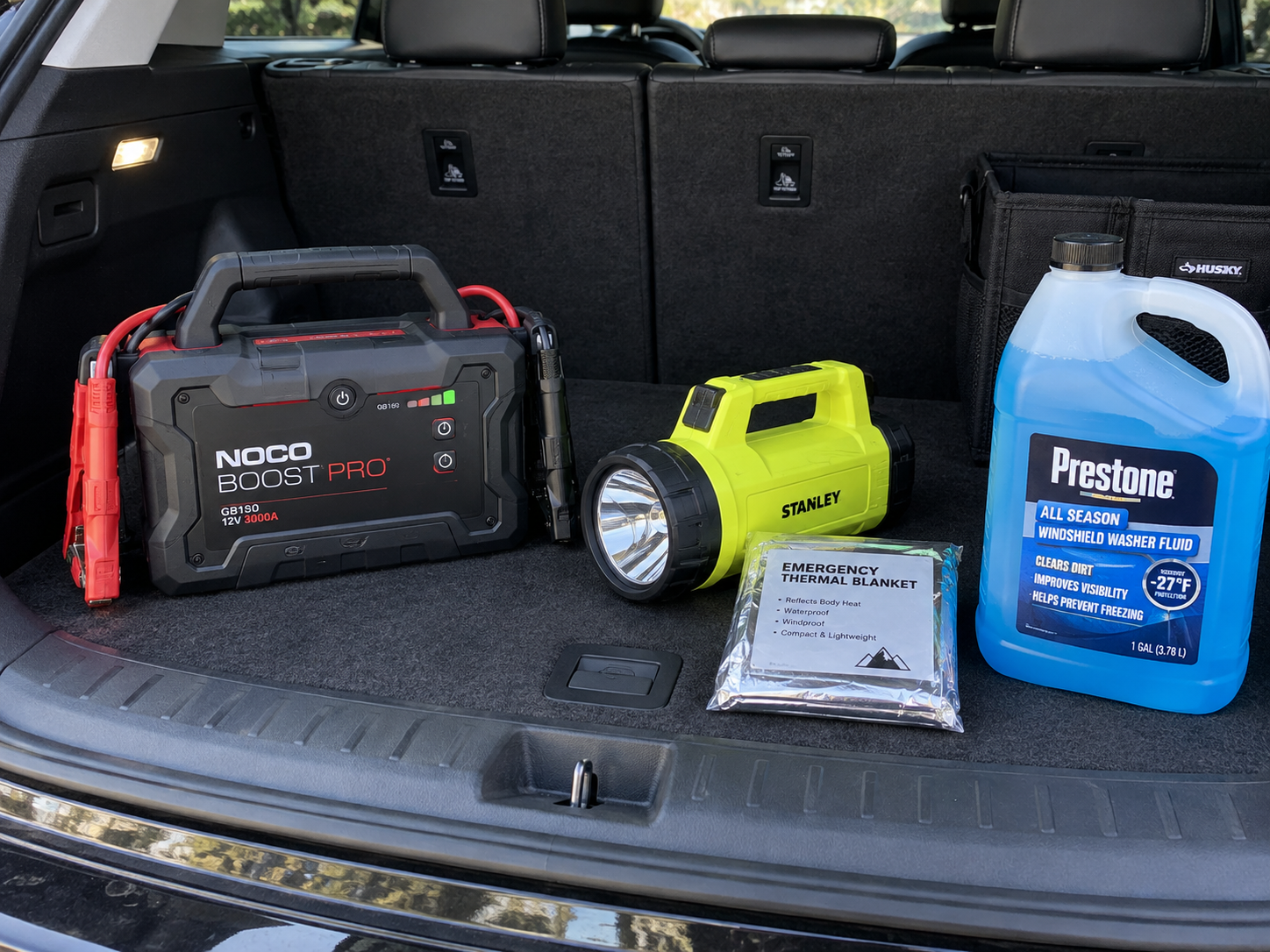 An emergency winter car trunk kit showing heavy-duty jumper cables, flashlight, thermal blanket, and windshield de-icer fluid
